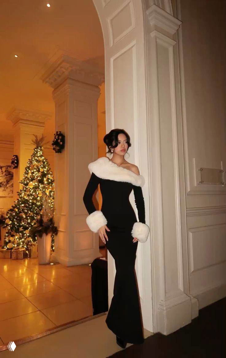 Модель в длином черном облегающем платье с белой меховой оторочкой на одном плече, на заднем плане рождественская ёлка и праздничный зал.