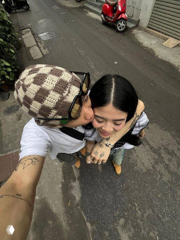 Пара делает селфи сверху: парень целует девушку в щёку на городской улице, видны татуировки на руке, скутер и асфальт.