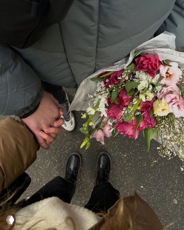 Крупный кадр рук пары, держащихся за руки рядом с букетом цветов; видны ботинки и тротуар — уличный романтический момент.