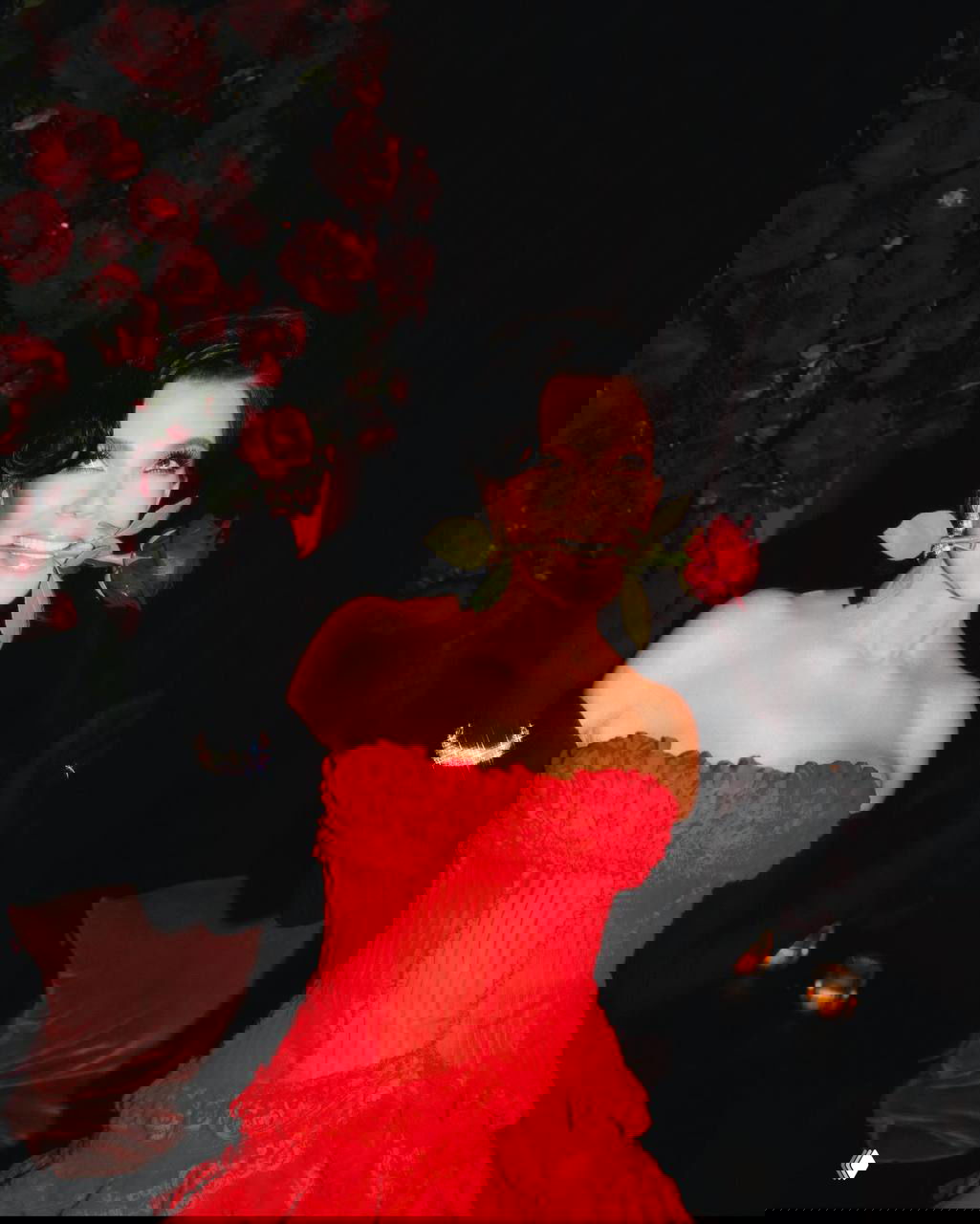 Крис Дженнер в роскошном красном многослойном платье и длинных черных перчатках, держит в зубах красную розу на фоне роз и свечей вечеринки.