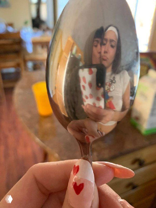 Отражение пары в ложке: девушка с маникюром в виде сердечек и парень видны в искажённом зеркальном отражении — бытовой романтический снимок.