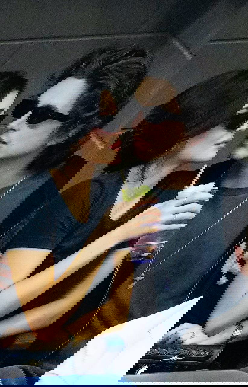 Двое молодых людей в тёмных очках сидят очень близко друг к другу; женщина держит стакан с напитком — кадр с публичного мероприятия на трибуне.