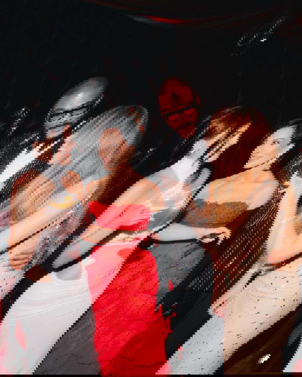 Кадр с Крис Дженнер в красном корсетном платье рядом с другими знаменитостями, все в вечерних нарядах, вокруг торжественная и камерная обстановка.