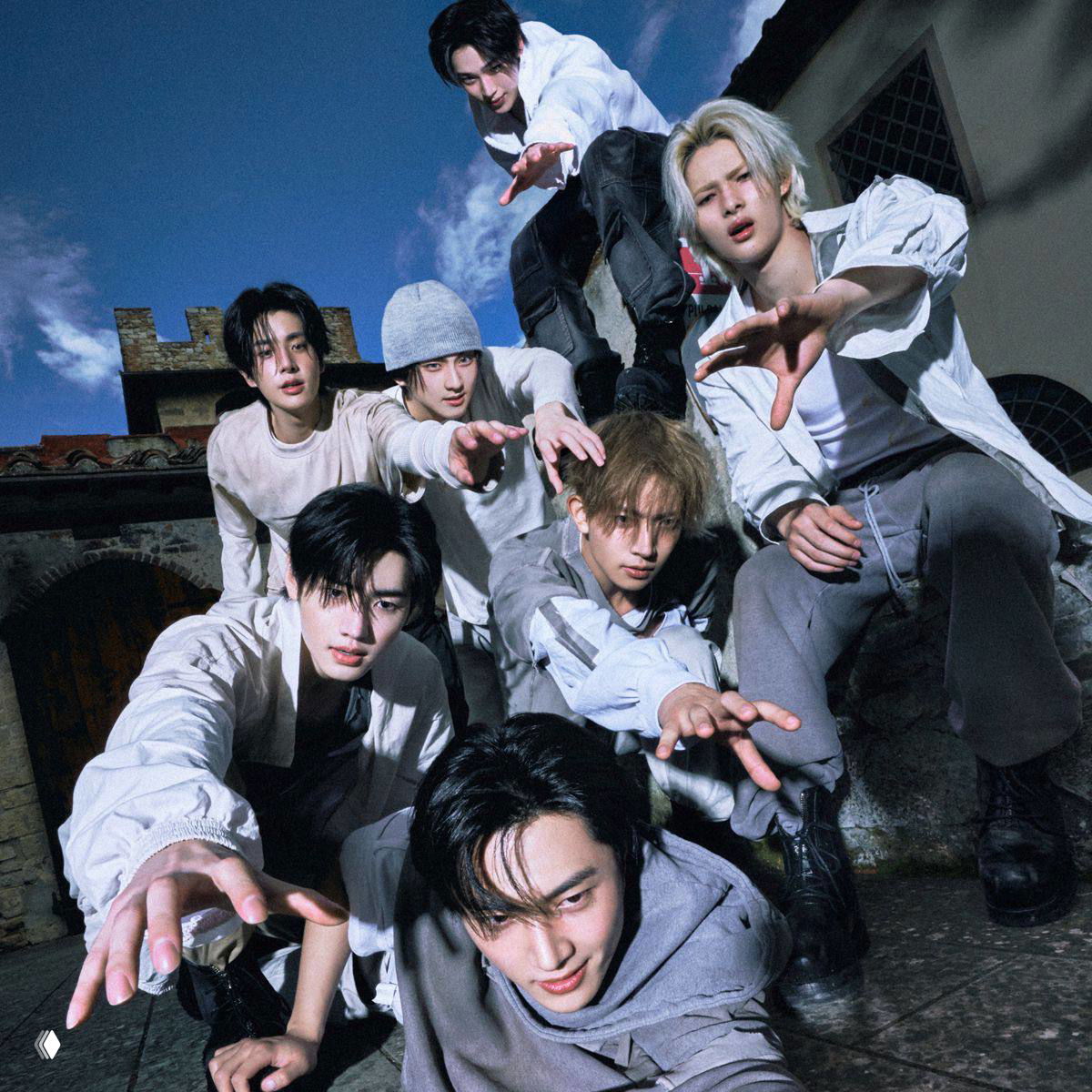 Промо‑фото k‑pop бойзбэнда на открытом воздухе: участники тянутся рукой к камере, кадр передаёт динамику и кинетическую энергию.