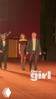 Ханде Эрчел выходит на сцену на премьере в платье, рядом сопровождающие, тёплая атмосфера и красный ковер; кадр сделан в зале показа.