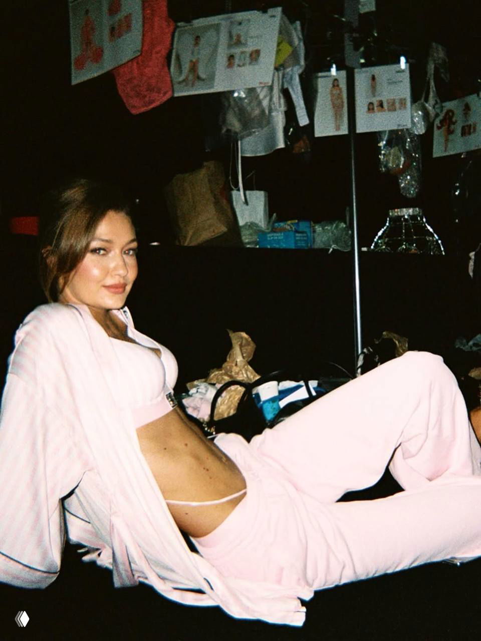 Модель в розовом комплекте отдыха и белья отдыхает за кулисами; на фото видны вешалки и аксессуары, атмосфера подготовки к показу.