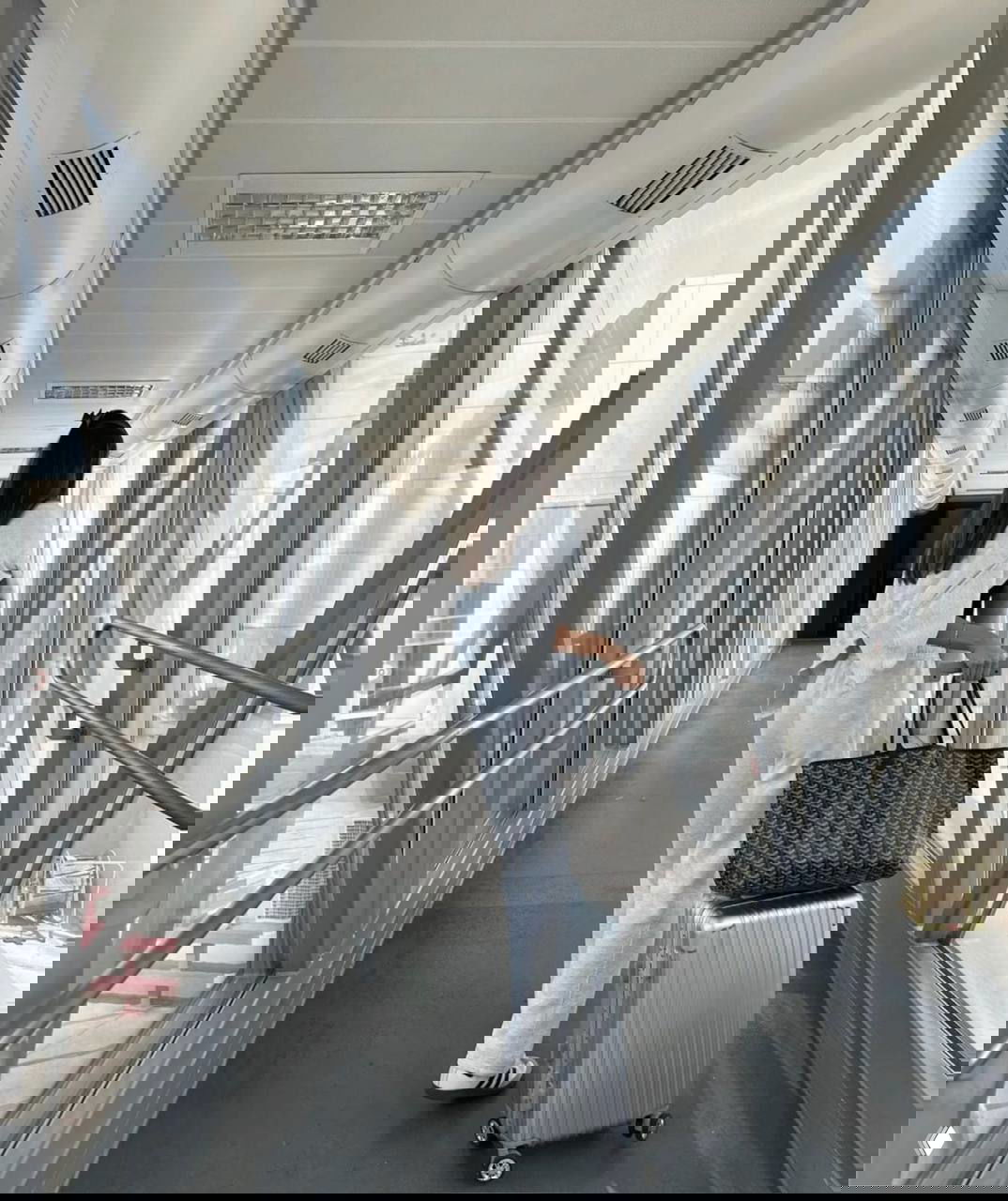 Девушка в светлом костюме с серебристым чемоданом и большой сумкой идёт по трапу аэропорта, вид сзади, дорожная сцена.