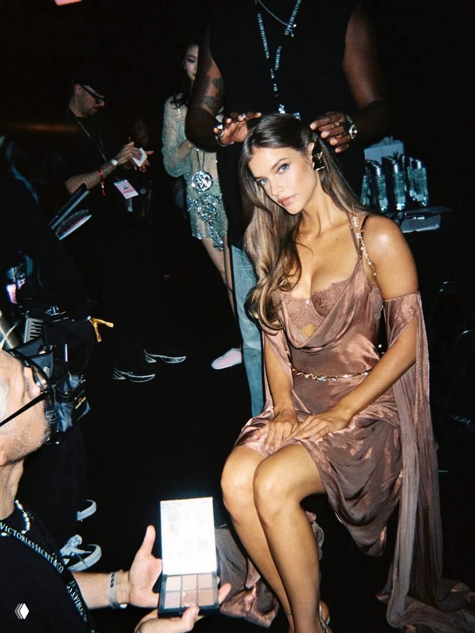 Модель в коричневом атласном платье сидит на стуле в окружении визажистов и ассистентов; кадр передаёт атмосферу подготовки к выходу на подиум.