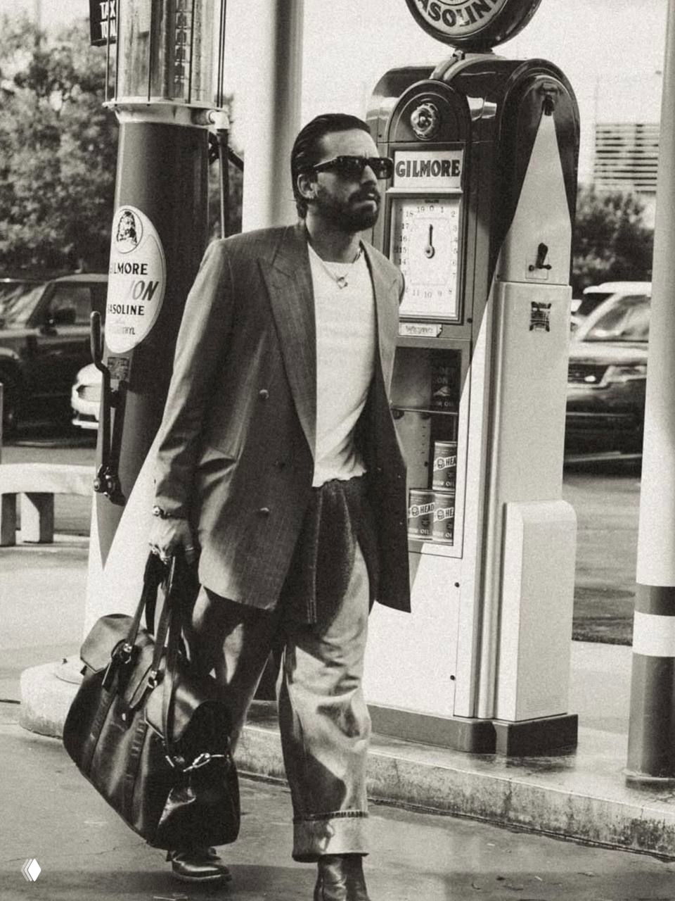 Чёрно-белая уличная фотография: мужчина в двубортном пальто и солнцезащитных очках, несёт дорожную сумку рядом с колонкой бензоколонки.