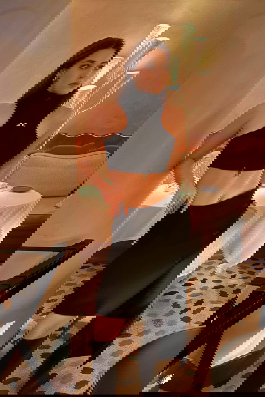 Девушка в ресторане в чёрном укороченном топе и асимметричной юбке, позирует у столика; интерьер с мягким светом и декоративной лампой.