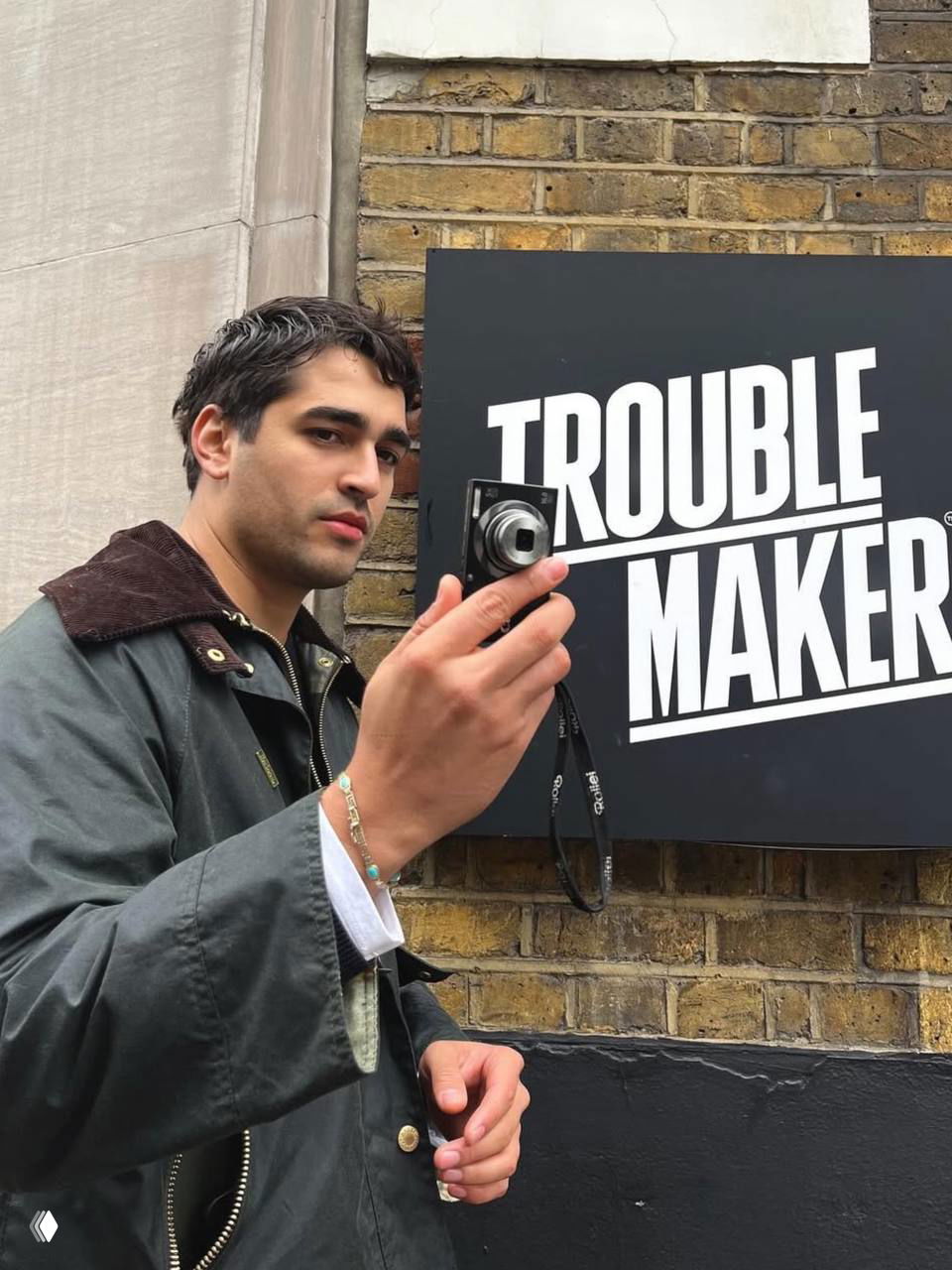 Мужчина на улице держит компактный фотоаппарат на фоне кирпичной стены и крупной рекламной вывески, повседневный уличный стиль и непринуждённая поза.