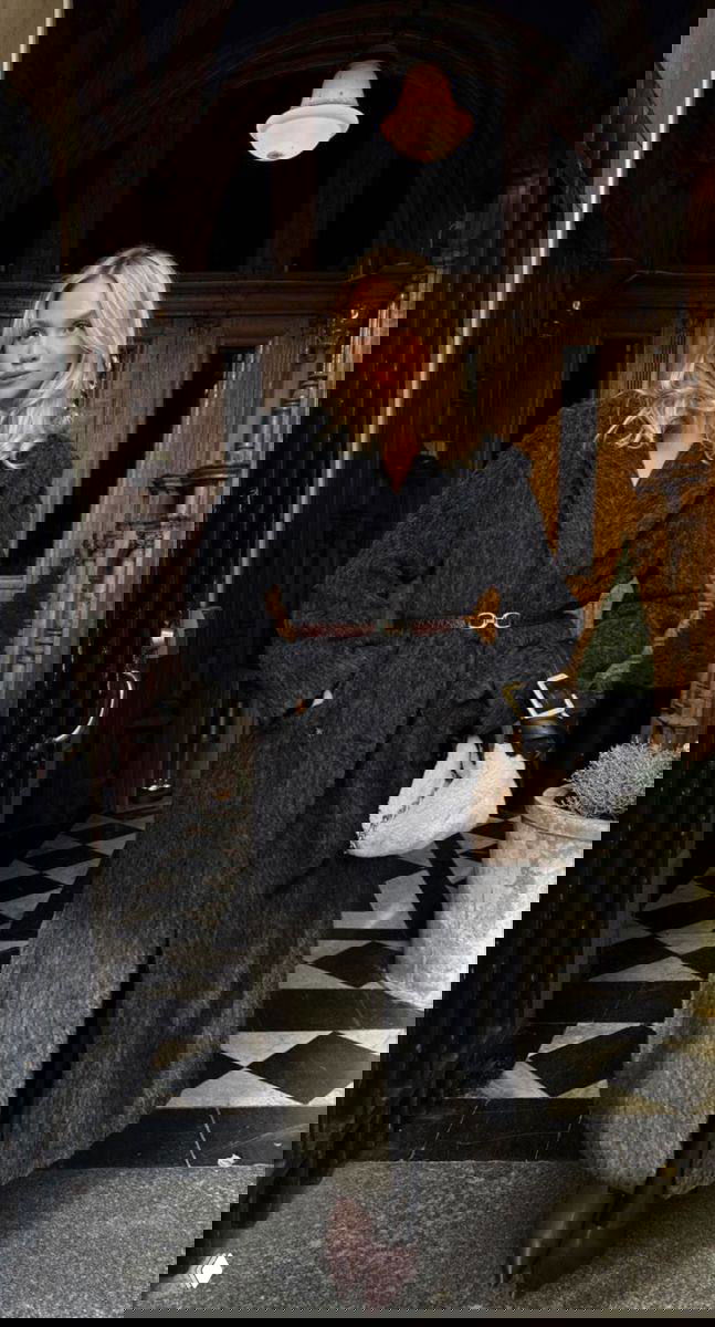 Женщина в тёмном пальто с поясом стоит в дверном проёме старинного здания, образ в уличном стиле, аксессуары заметны.