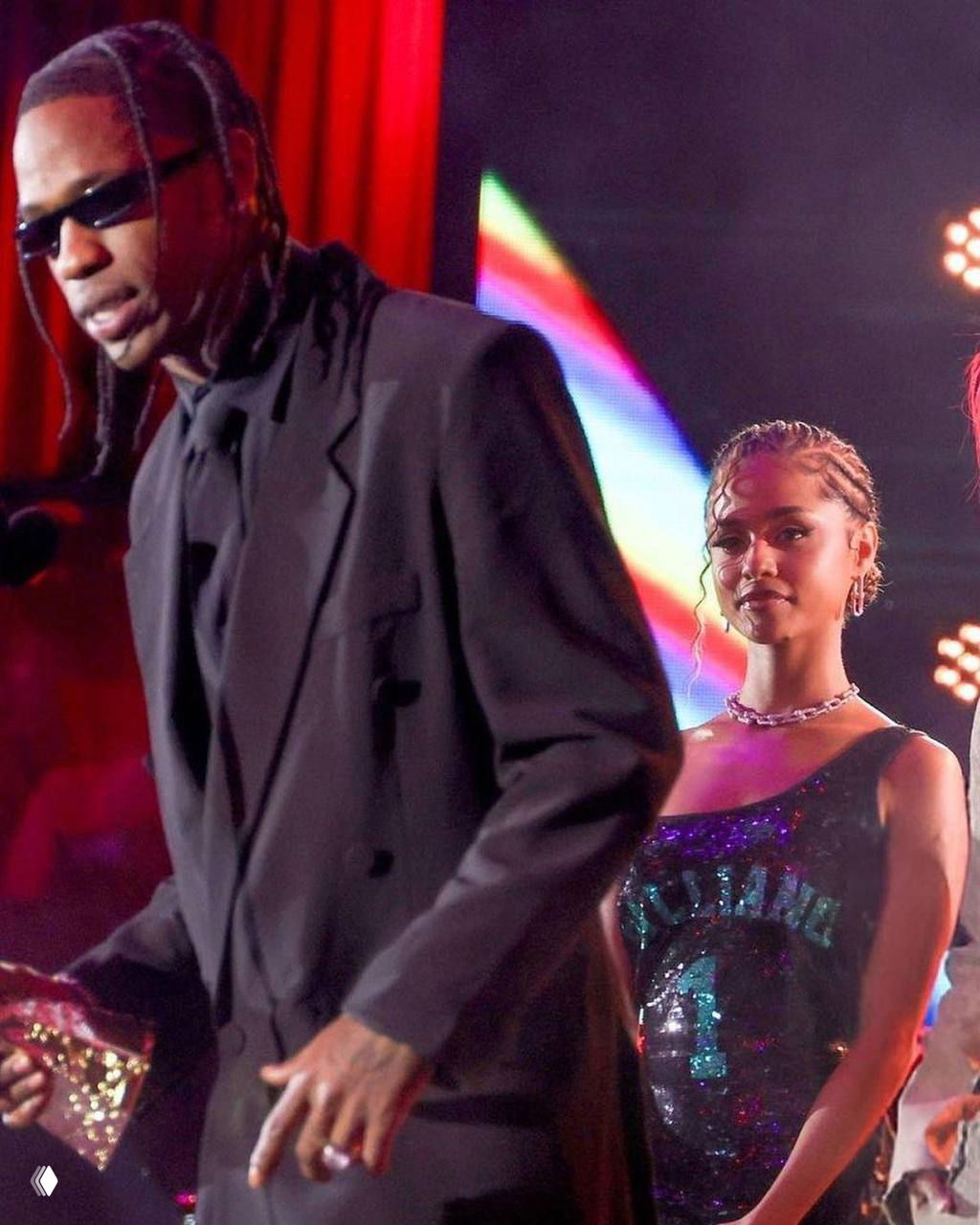 Трэвис Скотт в чёрном костюме выступает на сцене, Тайла стоит за ним в блестящем платье — сценические огни и публика вокруг.