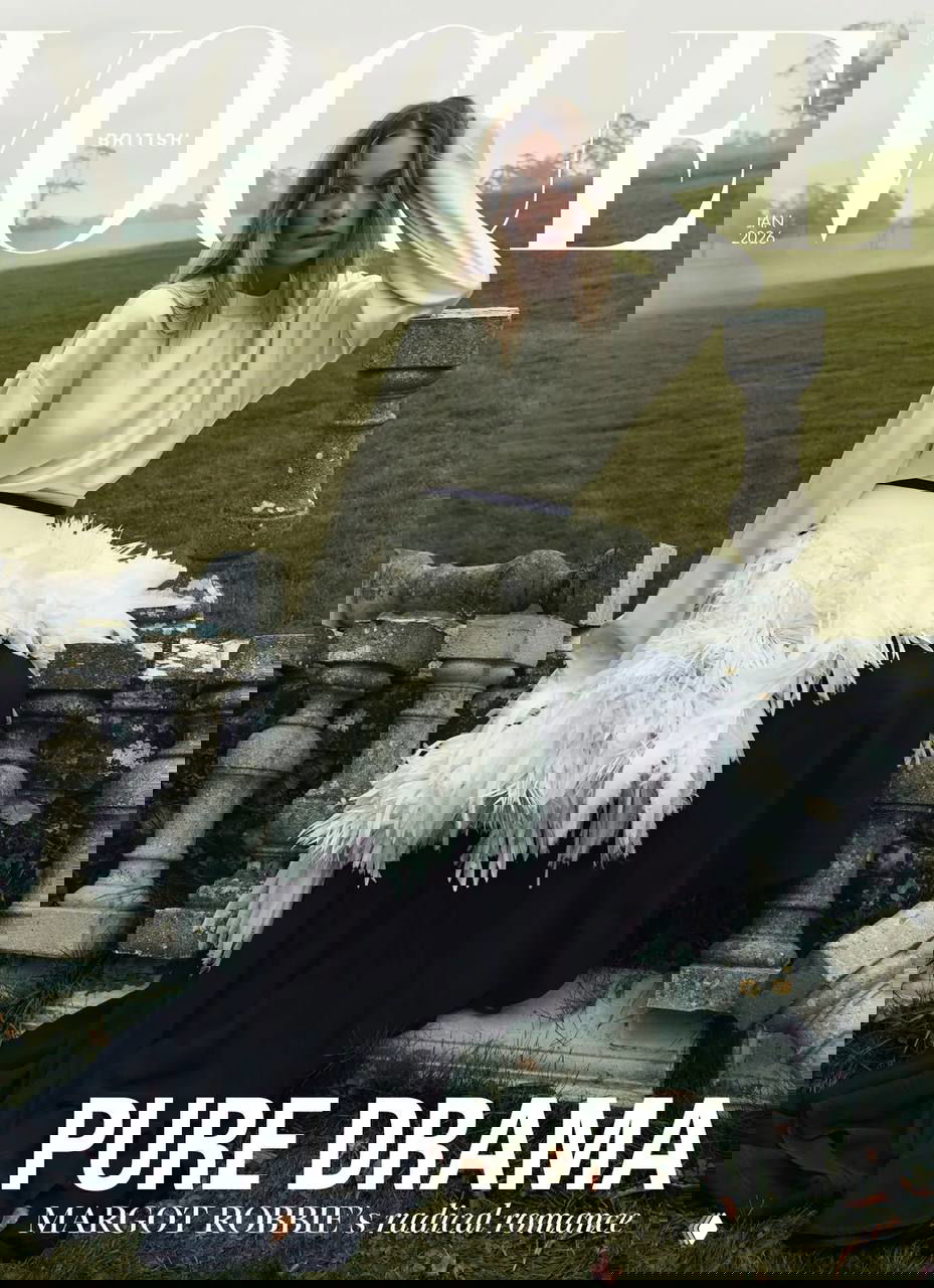 Женщина в длинном романтическом платье сидит на каменной ограде на фоне загородного пейзажа и туманного неба; кадр в стилистике модного журнала.