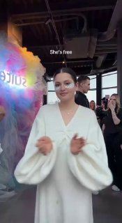 Момент с мероприятия: молодая женщина в белом наряде аплодирует на фоне яркой инсталляции с надписью «She's free», вокруг присутствуют посетители.