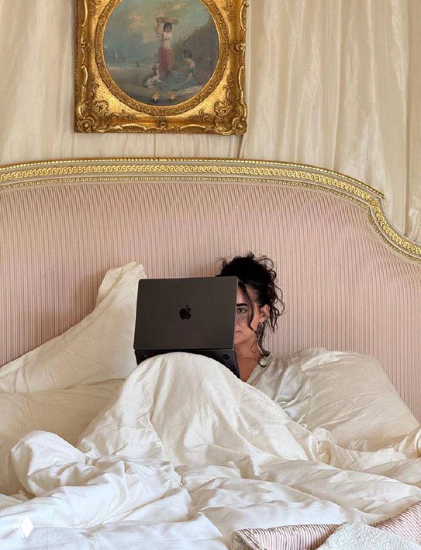 Человек в кровати с ноутбуком на фоне изысканного изголовья и мягкого текстиля — спокойная домашняя сцена в пастельных тонах.