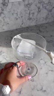 Рука держит стеклянный бокал с крупным кубиком льда на мраморной столешнице — кадр подготовки сервировки для холодного кофе.