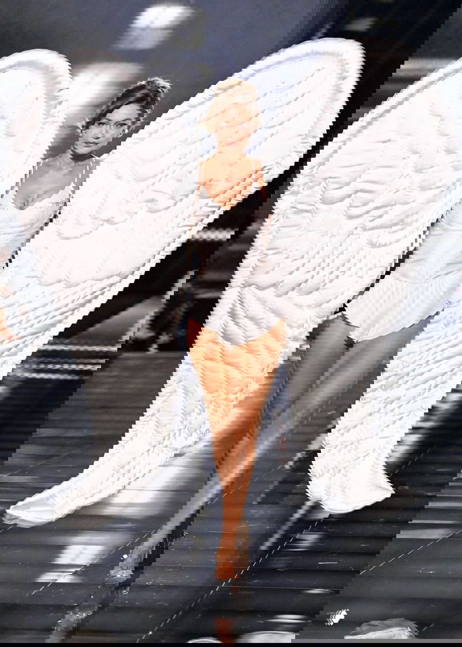 Модель на подиуме в белом корсете и больших мягких крыльях шоу Victoria’s Secret — полный рост, вид спереди, причёска в стиле «ракушка».