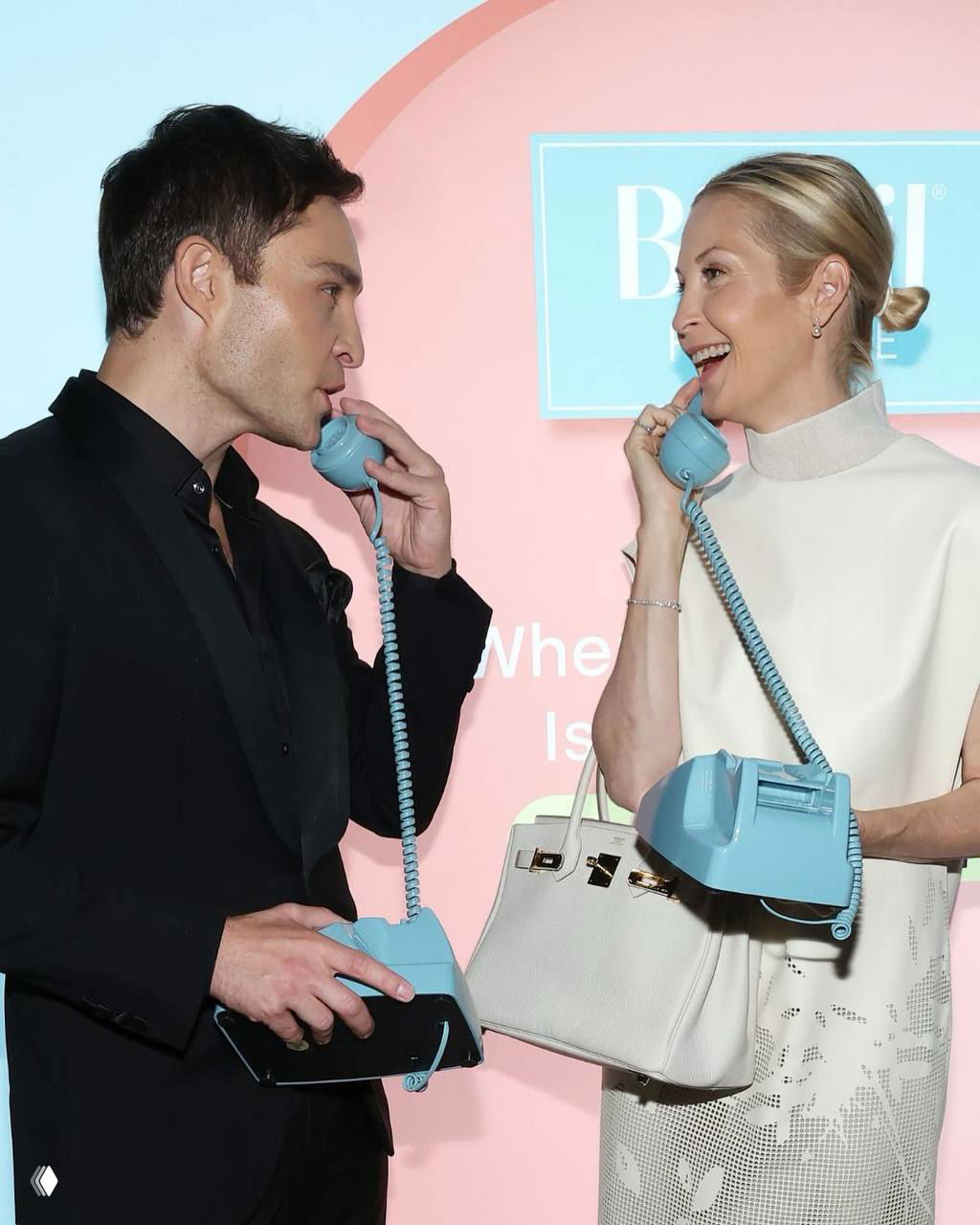 Эд Вествик и Келли Разерфорд на ужине Biosil: они улыбаются и держат голубые ретро-телефоны на фоне брендовой панели мероприятия.