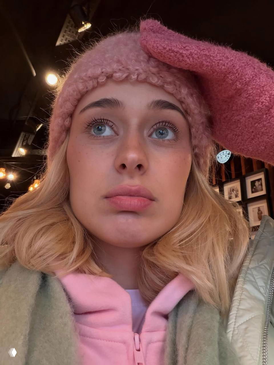 Крупный план лица Насти в розовой вязаной шапке и перчатке в кафе — естественный макияж, голубые глаза и задумчивый выразительный взгляд.