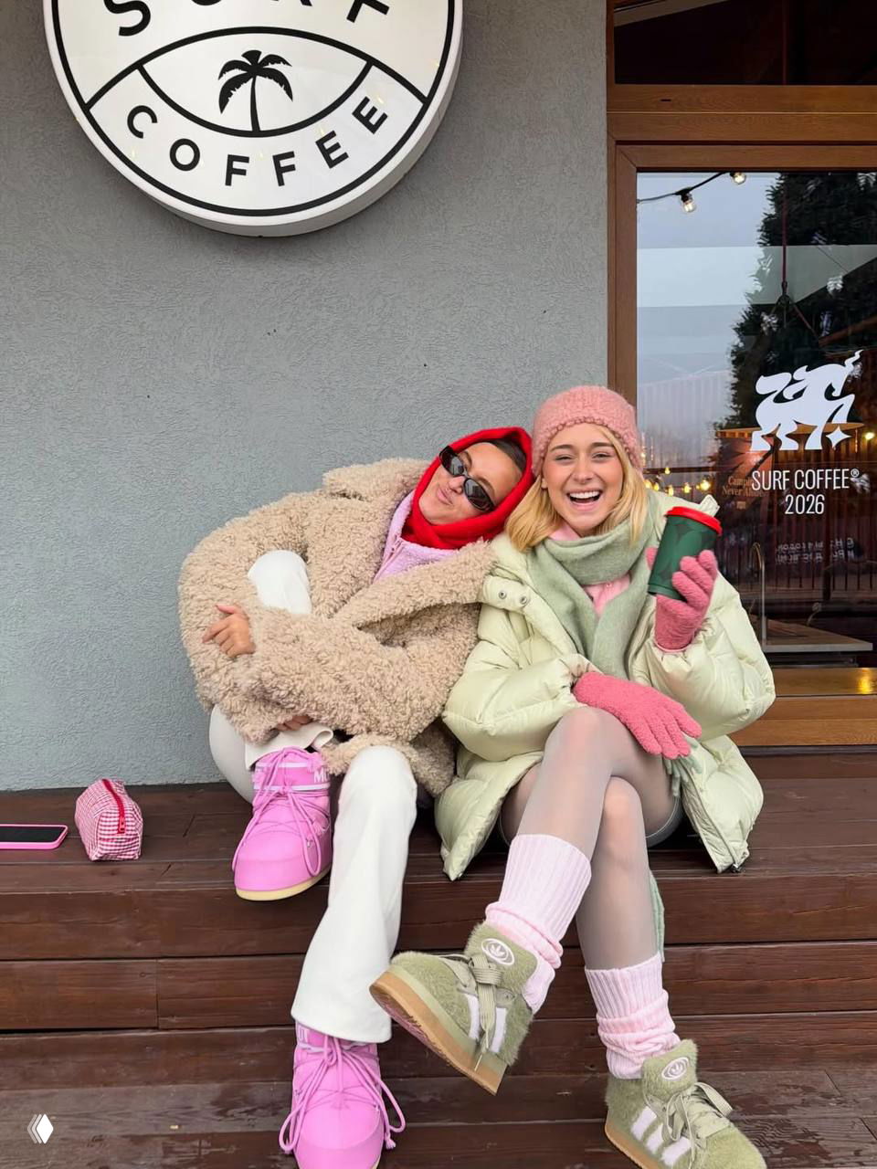 Настя и подруга сидят у входа в кафе Surf Coffee в тёплых куртках и ярких ботинках, смеются с чашками в руках на фоне вывески кофейни.