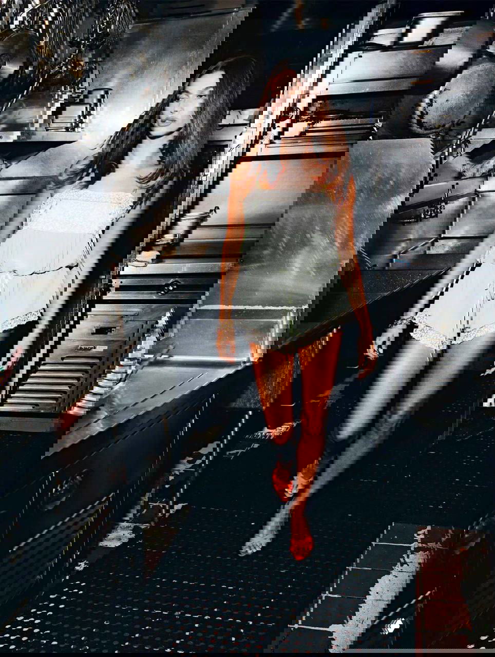 Фотография женщины, идущей по промышленной кухне в лёгком комбинезоне; контрастный фэшн‑кадр с повседневной утилитарной обстановкой.