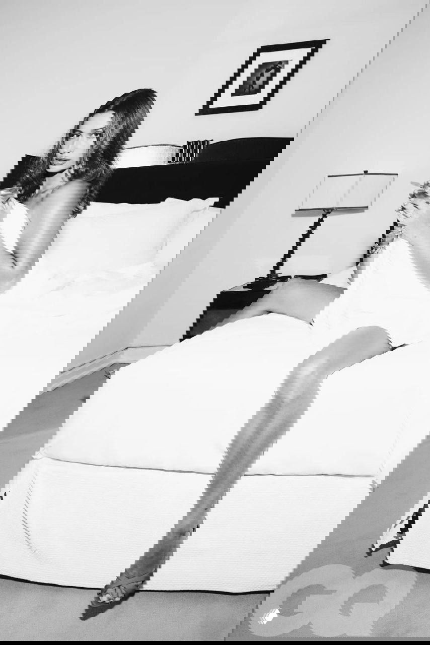 Чёрно‑белая фотография женщины, сидящей на краю кровати с бокалом в руке; элегантный журнальный портрет в интимной атмосфере.