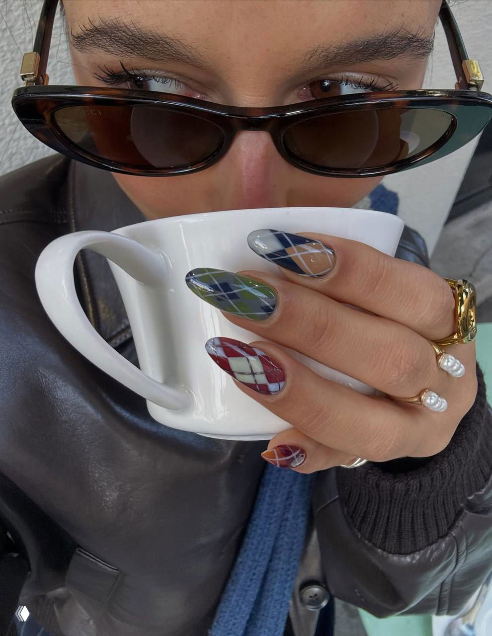 Крупный план руки с длинными ногтями в аргайл-узоре разных оттенков, держащей белую кружку; видны очки и кольца на пальцах.