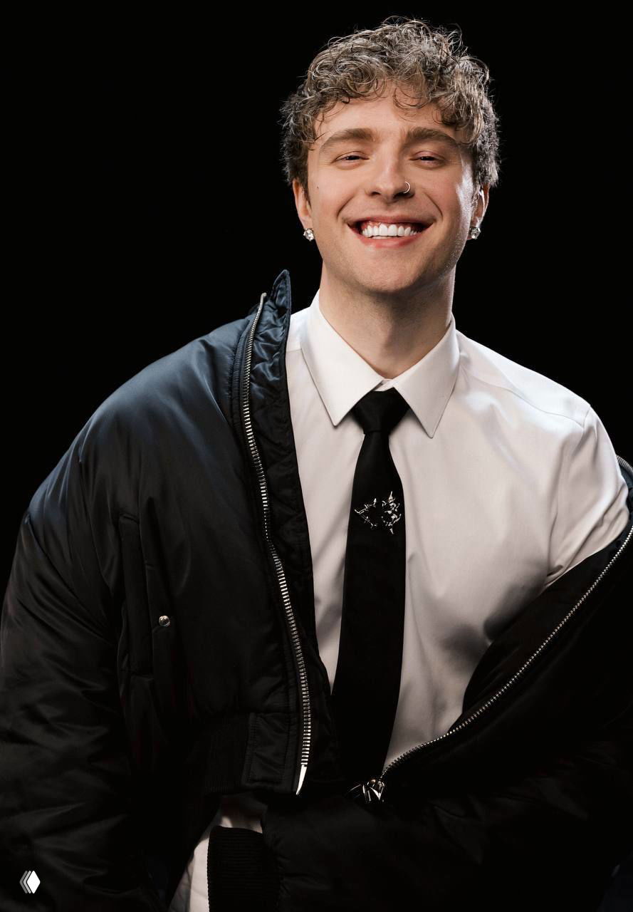 Портретный снимок улыбающегося молодого мужчины в белой рубашке, галстуке и чёрной куртке на тёмном студийном фоне.