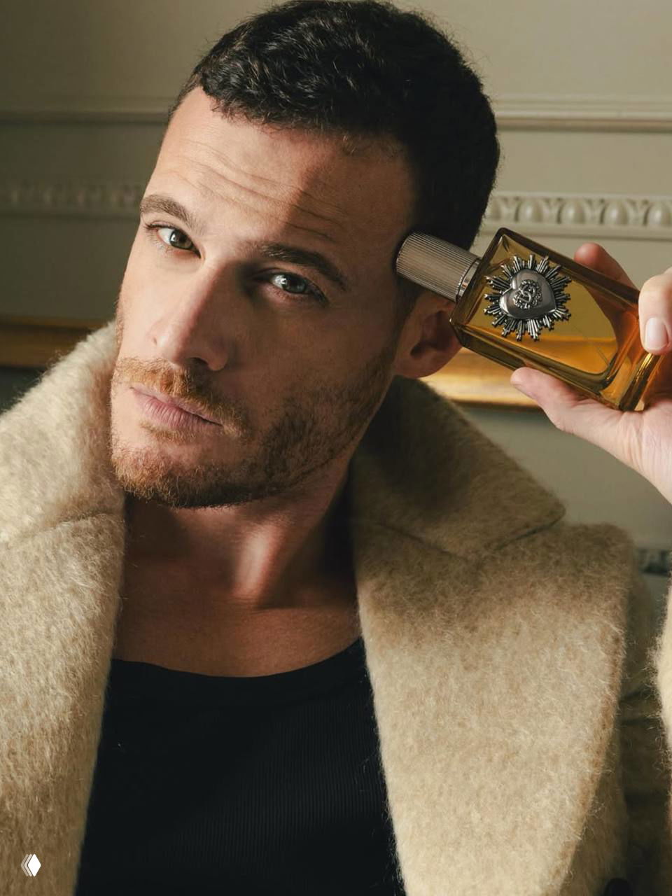 Крупный портрет Керема Бюрсина: мужчина в пальто держит флакон парфюма, выразительный взгляд и рекламная стилистика снимка.