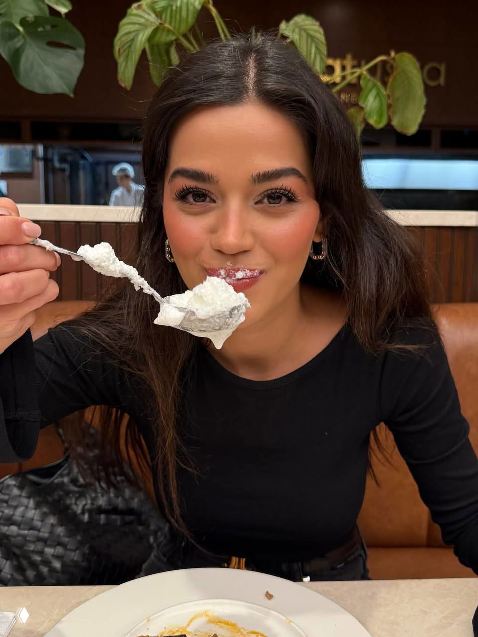 Селин Ягджиоглу улыбается за столом и ест десерт ложкой: тёмные длинные волосы, карие глаза и непринуждённая ресторанная сцена.
