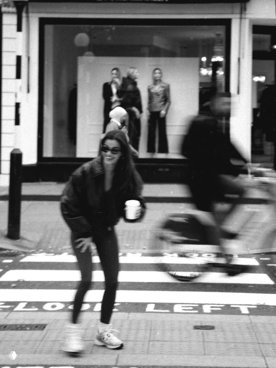 Чёрно‑белая уличная фотография: молодая женщина в солнцезащитных очках переходит пешеходный переход, размытый велосипедист на заднем плане.