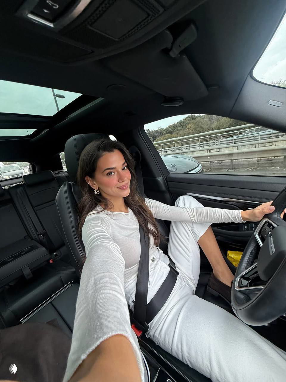 Селфи Селин Ягджиоглу в салоне автомобиля: она за рулём в ремне безопасности, расслабленный лайфстайл‑образ и городской фон за окном.