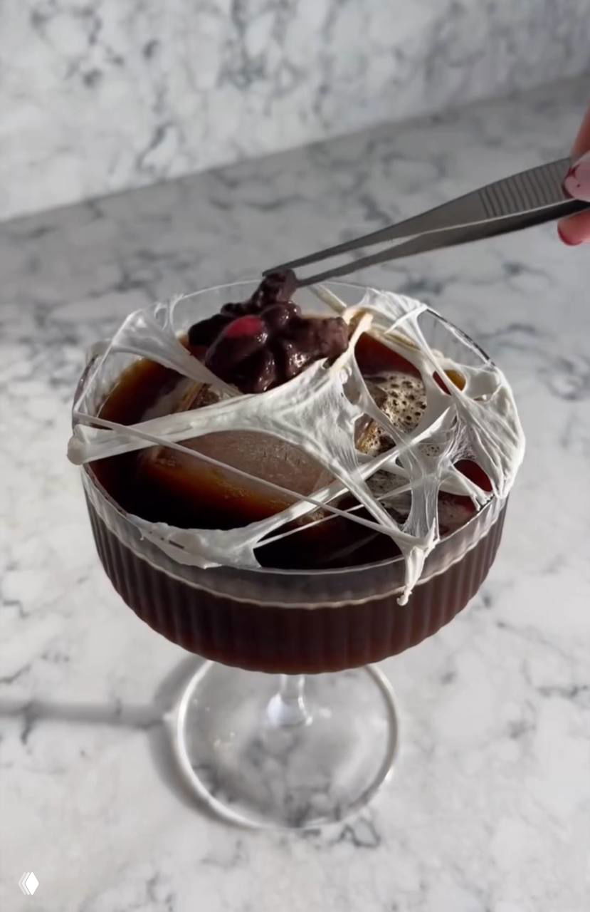 Крупный план готового напитка: холодный кофе в бокале, сверху натянута белая тянущаяся масса маршмеллоу в форме паутины.