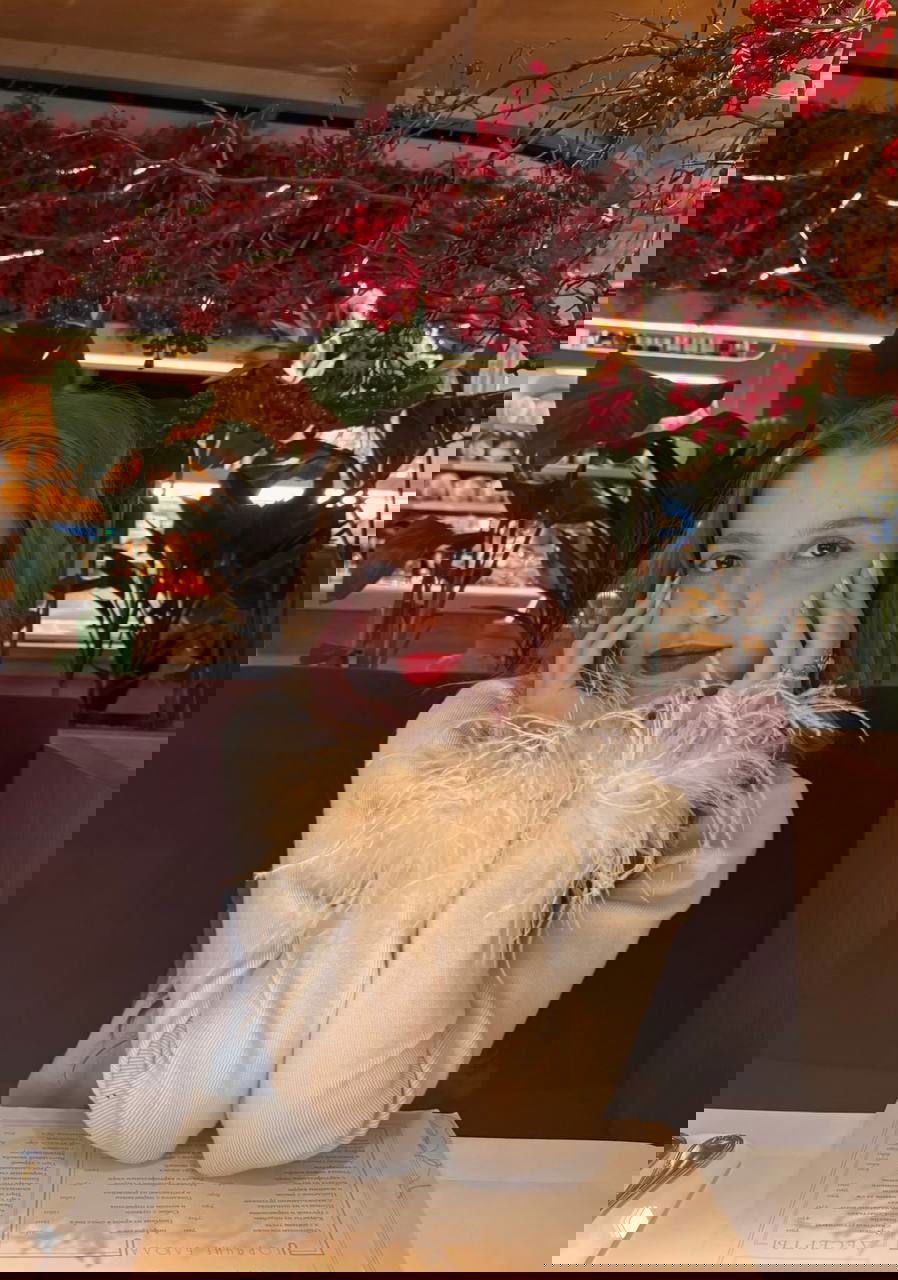 Девушка в кафе в светлом кардигане с перьевыми манжетами, красная помада и праздничный декор с ягодами на заднем плане.