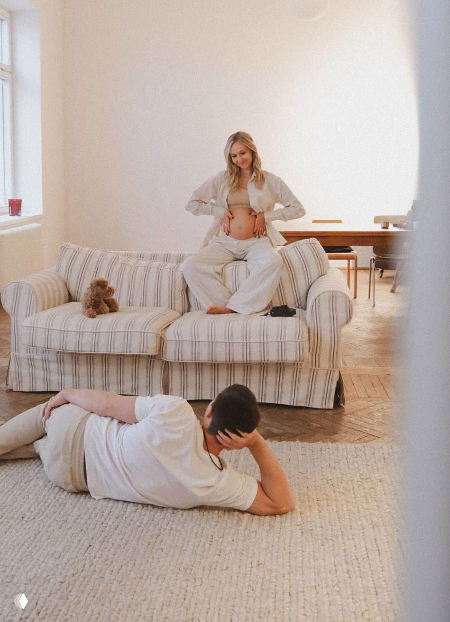 Широкий кадр в светлой комнате: женщина сидит на диване, позируя с видимым животом, мужчина лежит на полу и смотрит в её сторону — домашняя, спокойная сцена.