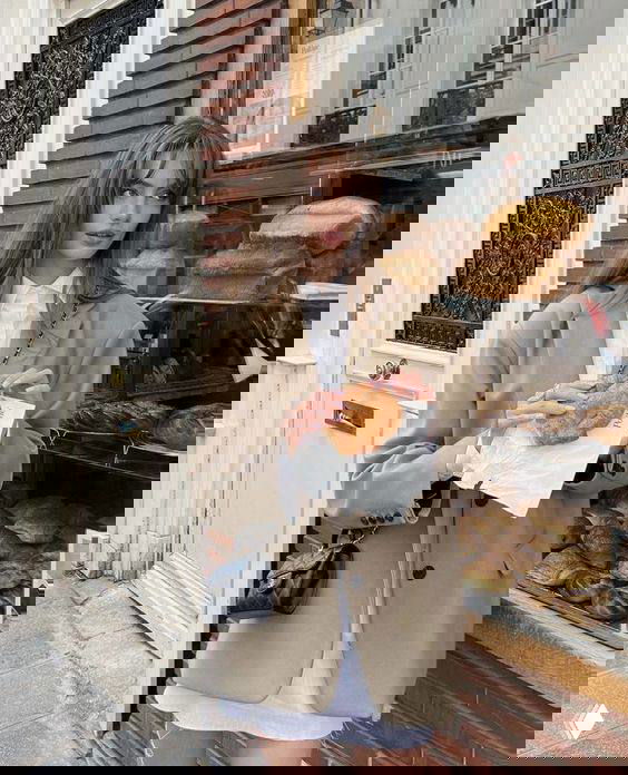 Молодая женщина в бежевом пиджаке у витрины пекарни в городском французском антураже держит круассан, уличная сцена и выпечка на фоне.