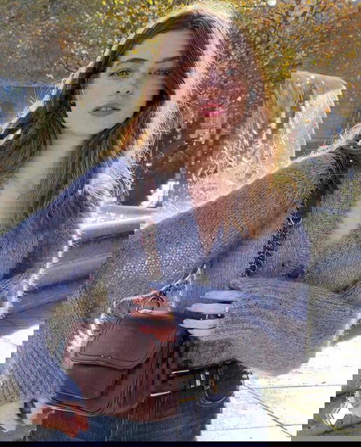 Девушка в мягком свитере на улице, естественная укладка волос и осеннее солнце создают лёгкую французскую эстетику с городским фоном.