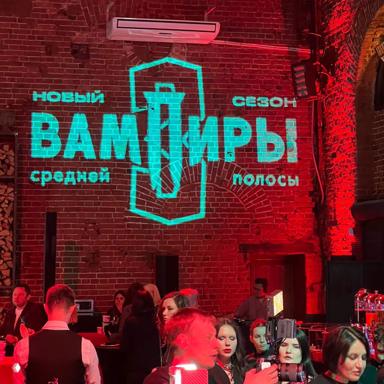 Проекция логотипа «Вампиры средней полосы» светится зелёным на кирпичной стене клуба, вокруг — гости и фотографы на мероприятии.