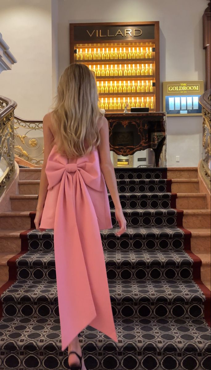 Женщина в розовом мини-платье с крупным декоративным бантом на спине, поднимается по богато отделанной лестнице в интерьере отеля.