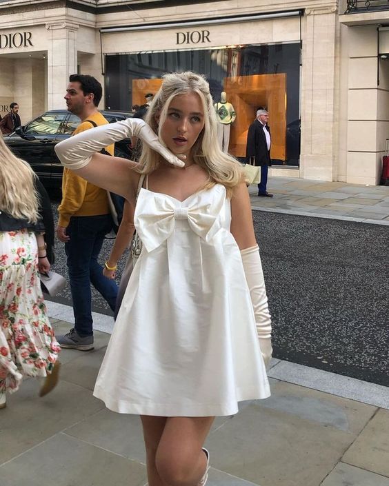 Молодая женщина в белом коротком платье с крупным бантом спереди идёт по улице рядом с витриной Dior, городской фэшн-образ.