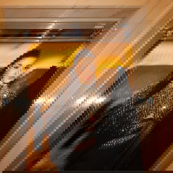 Женщина в блестящем вечернем наряде с крупными пайетками стоит у стеклянной двери или лифта, отражения и тёплый свет создают эффект праздника.