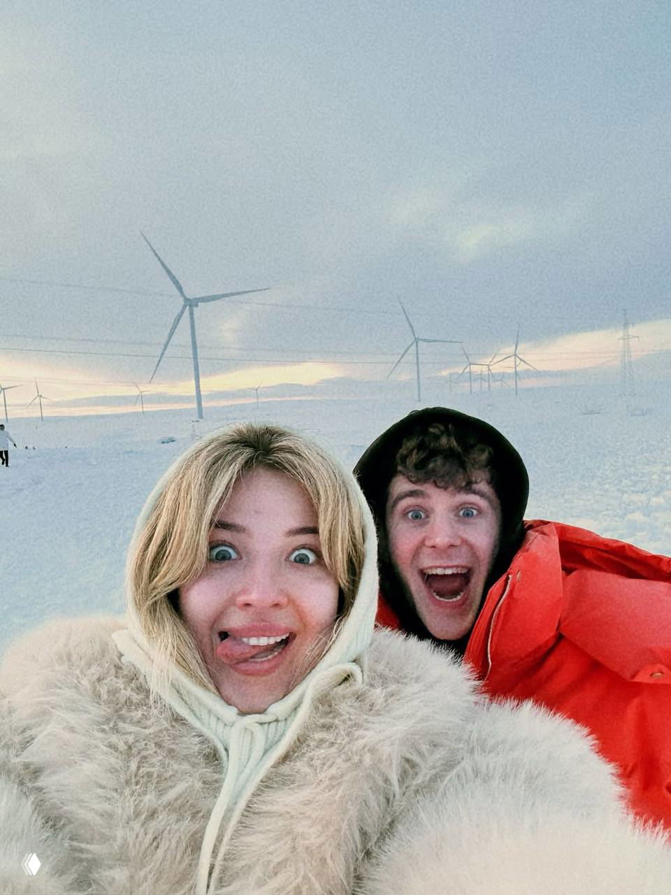 Селфи Ани Покров и Артура Бабича на заснеженном поле с видимыми ветряными турбинами на фоне; пара в теплой одежде, улыбается в холоде