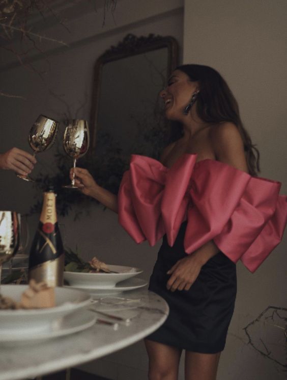 Женщина в чёрной юбке и ярком розовом лифе с объёмным бантом сидит за столом с бокалами и шампанским, праздничная атмосфера ужина.