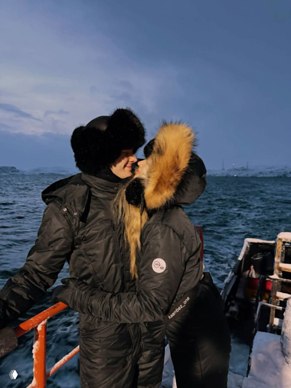 Аня Покров и Артур Бабич обнимаются и целуются на палубе судна у заснеженного моря; пара в темных зимних пуховиках с меховыми капюшонами