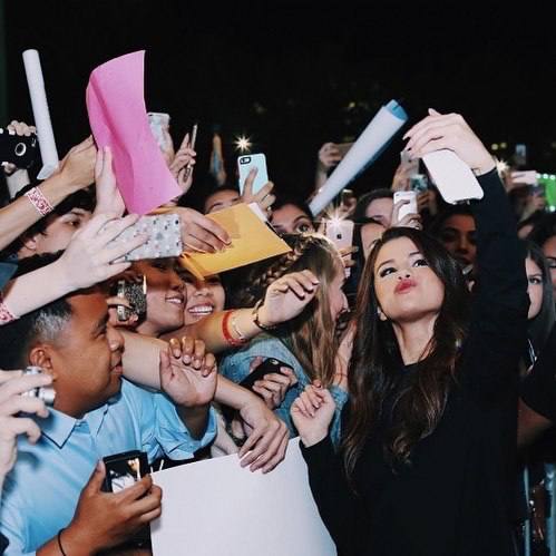 Ночная сцена у ограждения: женщина делает селфи или позирует перед толпой фанатов, люди протягивают листы бумаги и телефоны для автографа.
