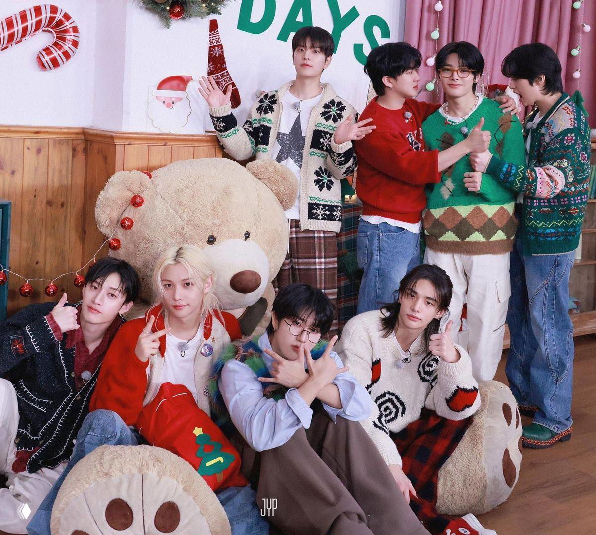 Stray Kids на групповой фотографии у большого плюшевого медведя, участники в уютных свитерах и праздничном декоре, тёплый предновогодний кадр