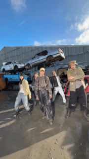 Группа из нескольких участников в уличной локации на фоне стопки автомобилей; коллективный кадр в стиле клипа с уличным антуражем.