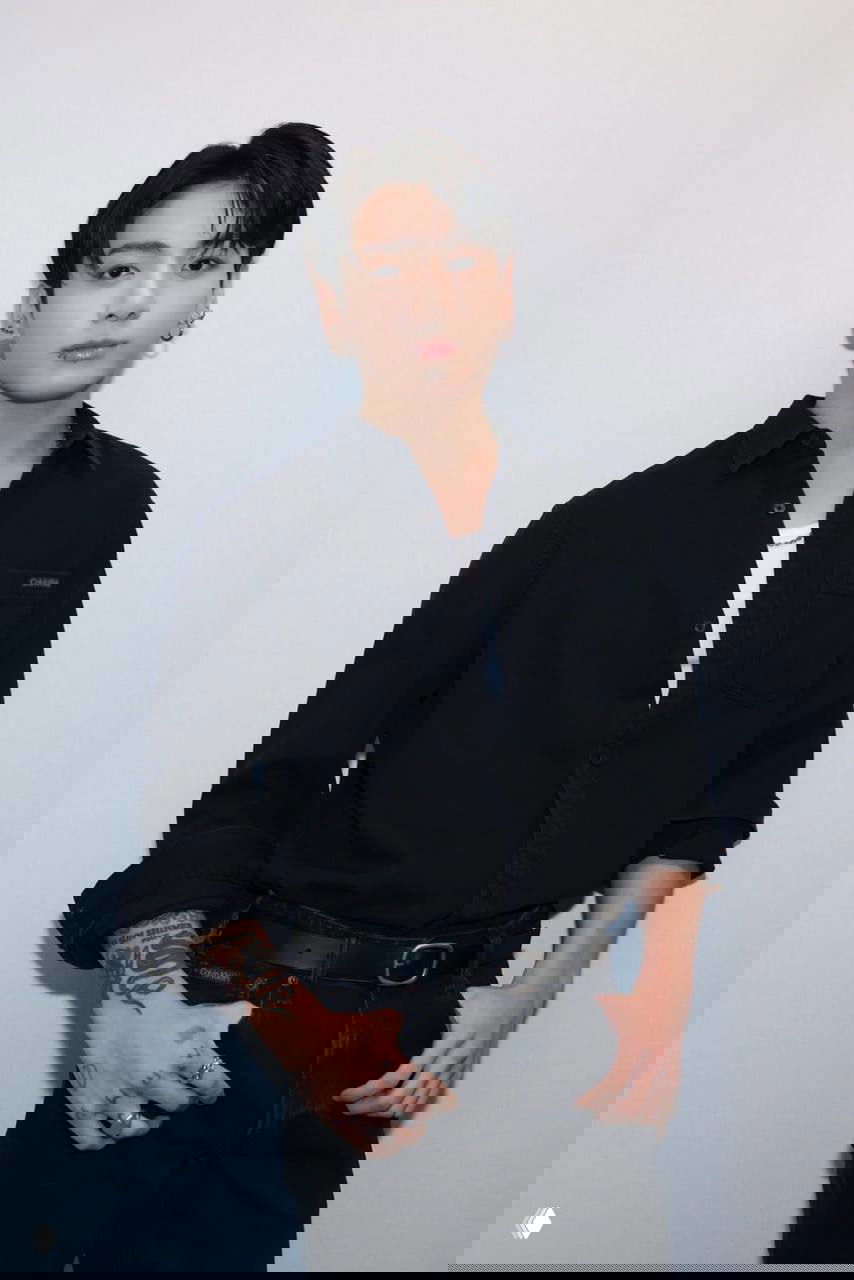 Фото молодого человека в чёрной рубашке и джинсах с тату и украшениями, позирующего на светлом фоне — портрет в стиле K‑pop.