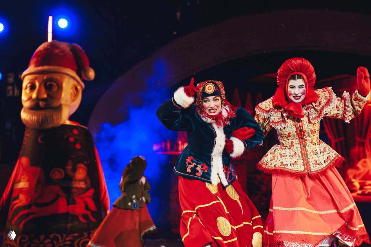 Два улыбающихся актёра в ярких костюмах вблизи сцены, вокруг — элементы народного орнамента и сценография в русском стиле.